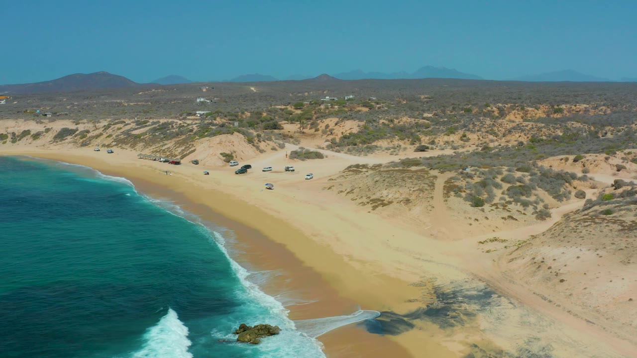 High aerial frame over Shipwrecks Los Cabos with blue-green ocean curving into the left foreground, a foamy wave near a rock, broad sand in the center, low dunes and parked vehicles inland, and no visible people.