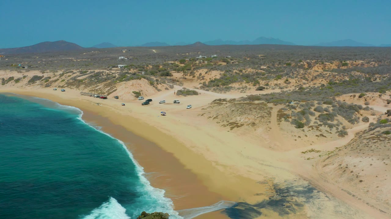 Mid-clip aerial frame at Shipwrecks Los Cabos with the ocean pressed along the left edge, a wide band of wet sand on the beach, dune tracks and scrub inland, parked vehicles near the upper beach, and no visible people.