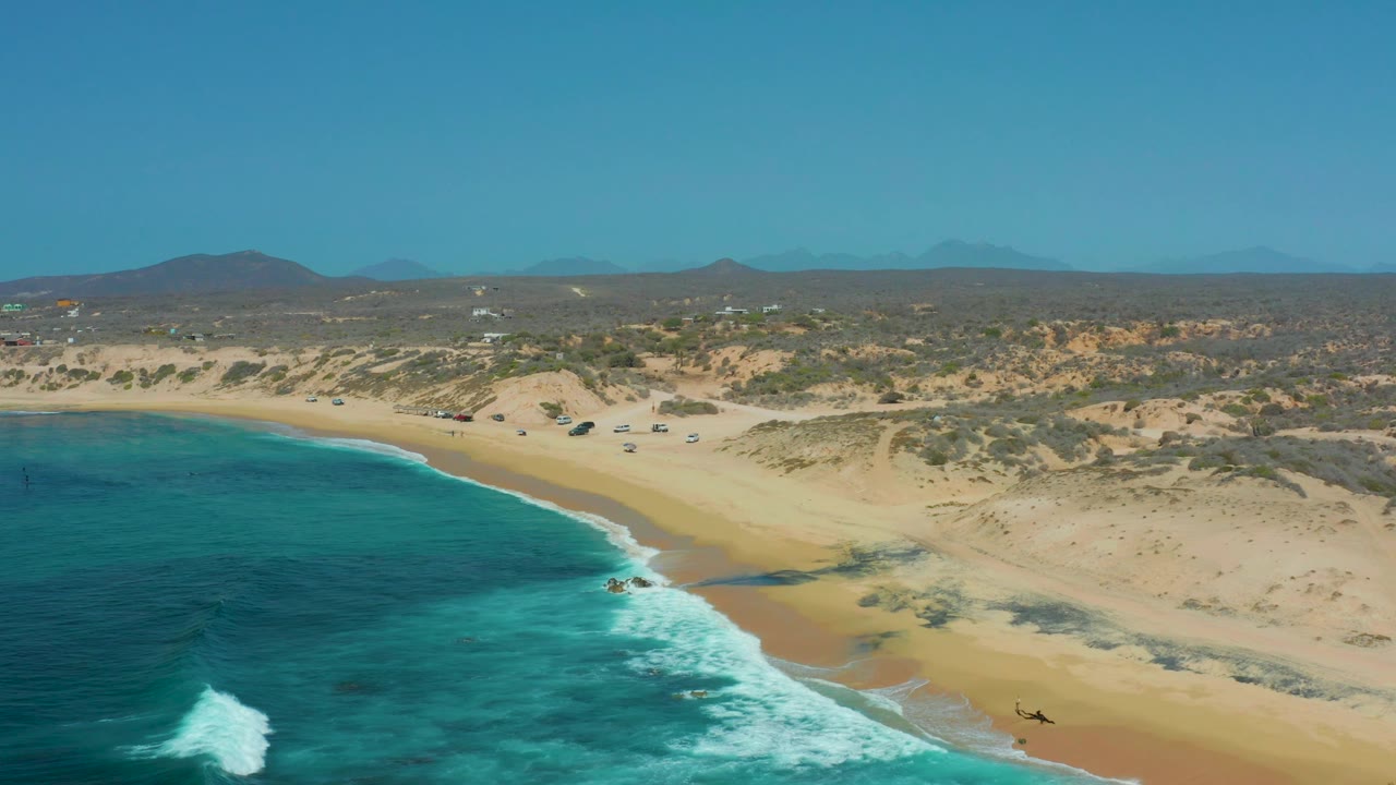 Aerial frame over Shipwrecks Los Cabos showing blue water at left, white surf on a sandy shore, low dunes and scrub inland, and no visible people.