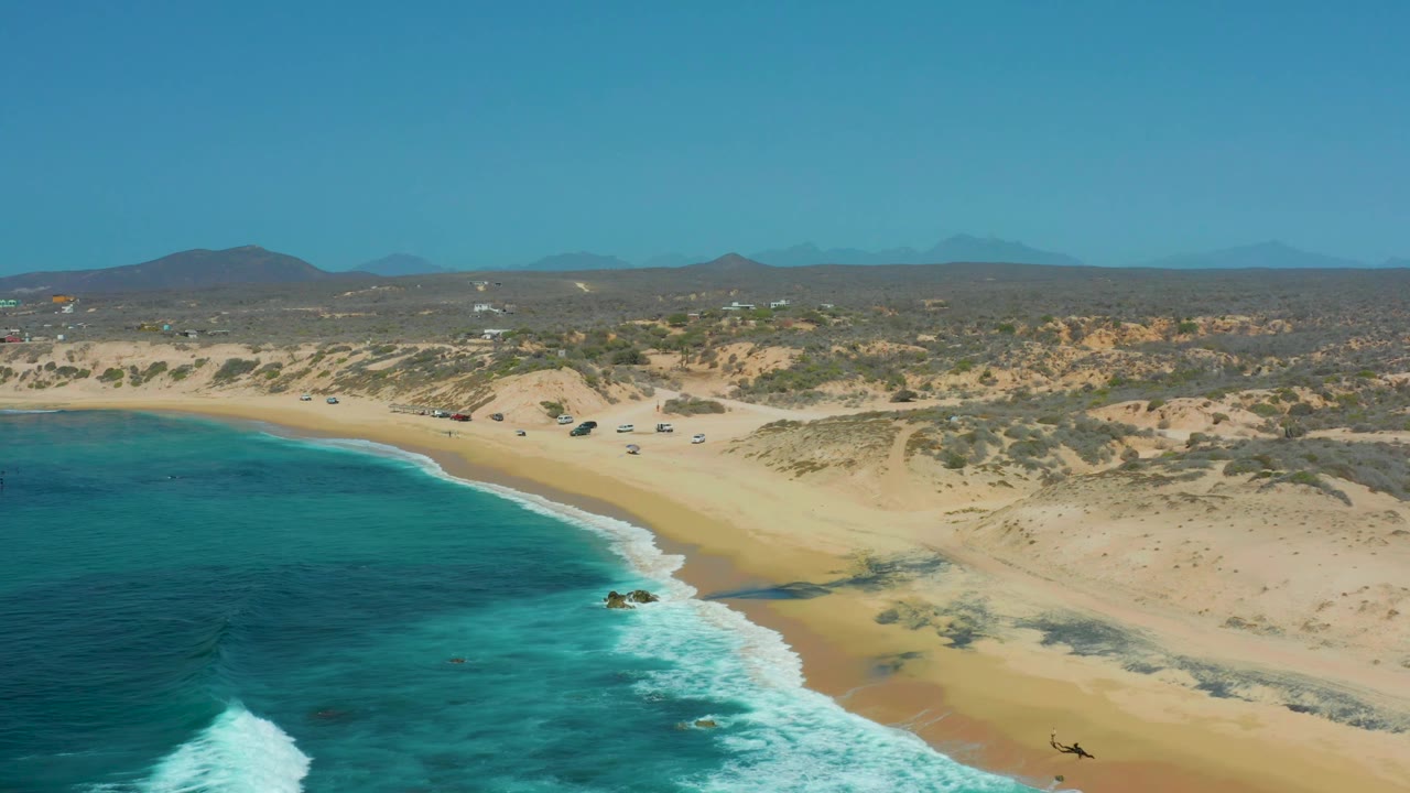 Shipwrecks Los Cabos shoreline seen from above, with turquoise water pressing into the lower left, a pale beach curving across frame, dunes inland, and no visible people.
