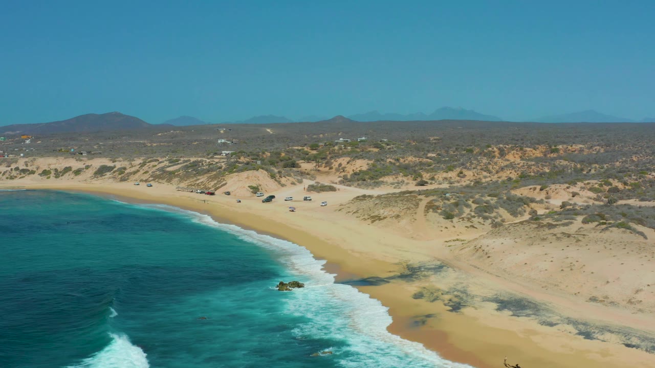 Wide aerial beach view with blue ocean, breaking surf, sandy dunes, scrub, and hills.