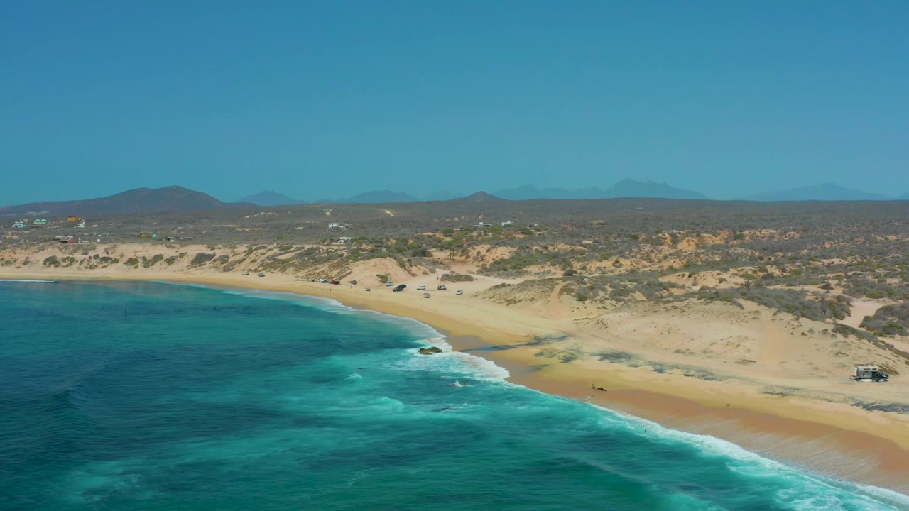 Shipwrecks Los Cabos shoreline one second in, with foamy waves tracing the beach, pale dunes behind the sand, small distant vehicles near the dune line, and no visible people.