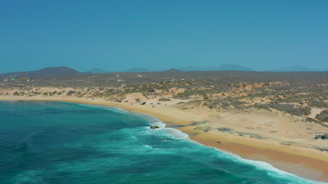 Aerial view over Shipwrecks Los Cabos showing the beach curve more fully, with turquoise water filling the left foreground, waves breaking near shore, dune scrub in back, and no visible people.