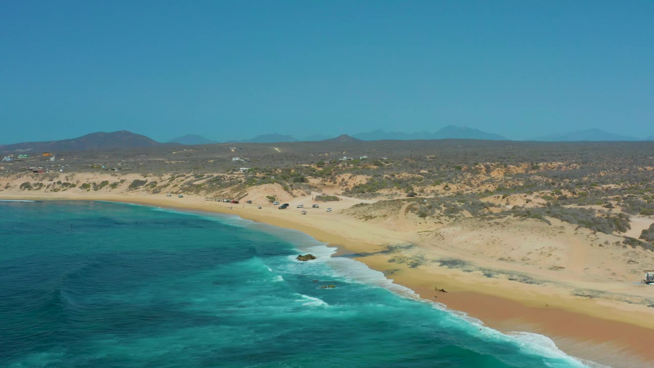 Mid-clip aerial frame at Shipwrecks Los Cabos with more open water in the foreground, white surf bands reaching the sand, low dune ridges beyond the beach, and no visible people.