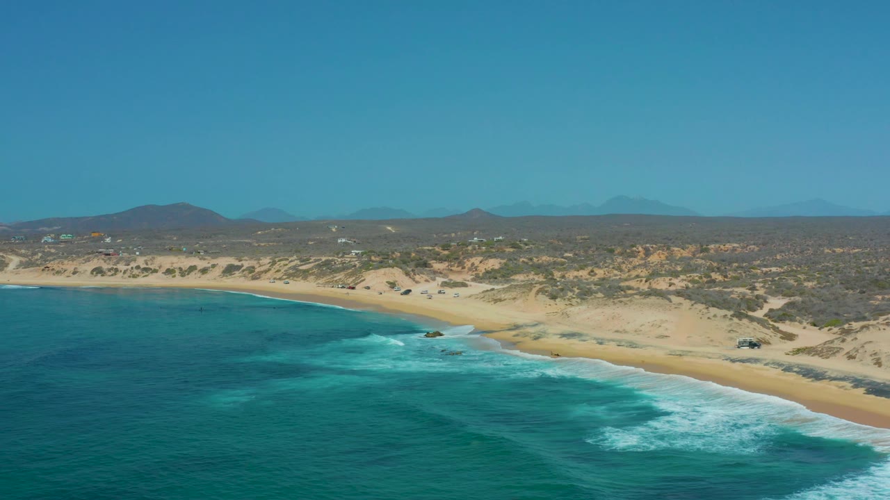 At 0:01, Shipwrecks Los Cabos appears from an elevated angle as foamy waves spread across turquoise water toward a broad sand beach, with scrub dunes and no visible people.