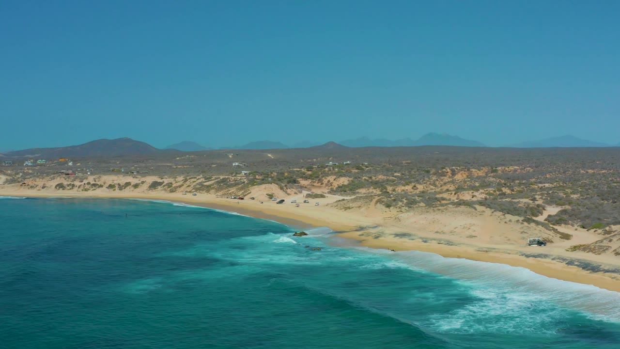 At 0:03, the frame holds a wider band of turquoise water in the foreground, small breaking waves at the shore, sandy dunes across the middle distance, distant hills, and no visible people.