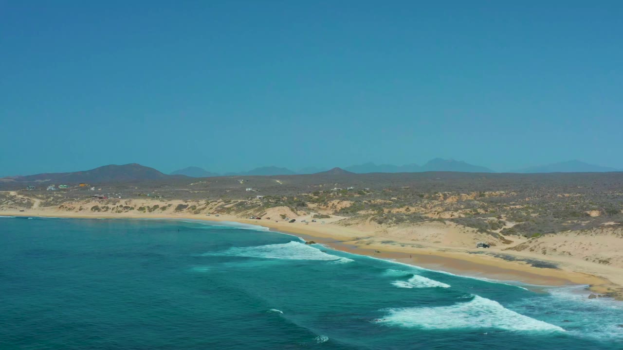 Opening frame over Shipwrecks Los Cabos shows turquoise water, white surf, a sandy shoreline, dune scrub, distant hills, clear sky, and no visible people.