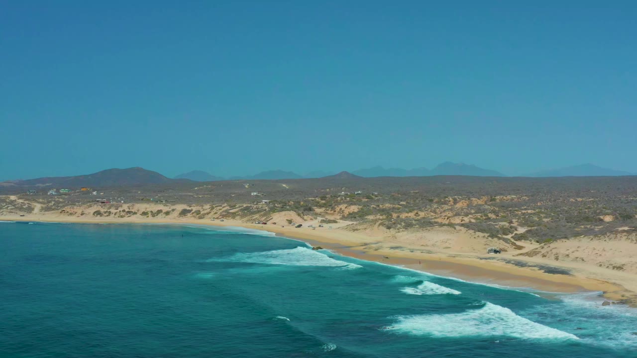At 0:01, Shipwrecks Los Cabos appears from a high coastal angle as surf lines move toward the sand, with scrub dunes and low hills under a clear sky and no visible people.