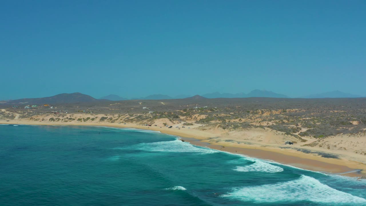 At 0:03, Shipwrecks Los Cabos is framed with the ocean filling the left and lower areas, a long sandy beach to the right, pale dunes behind it, distant hills, and no visible people.