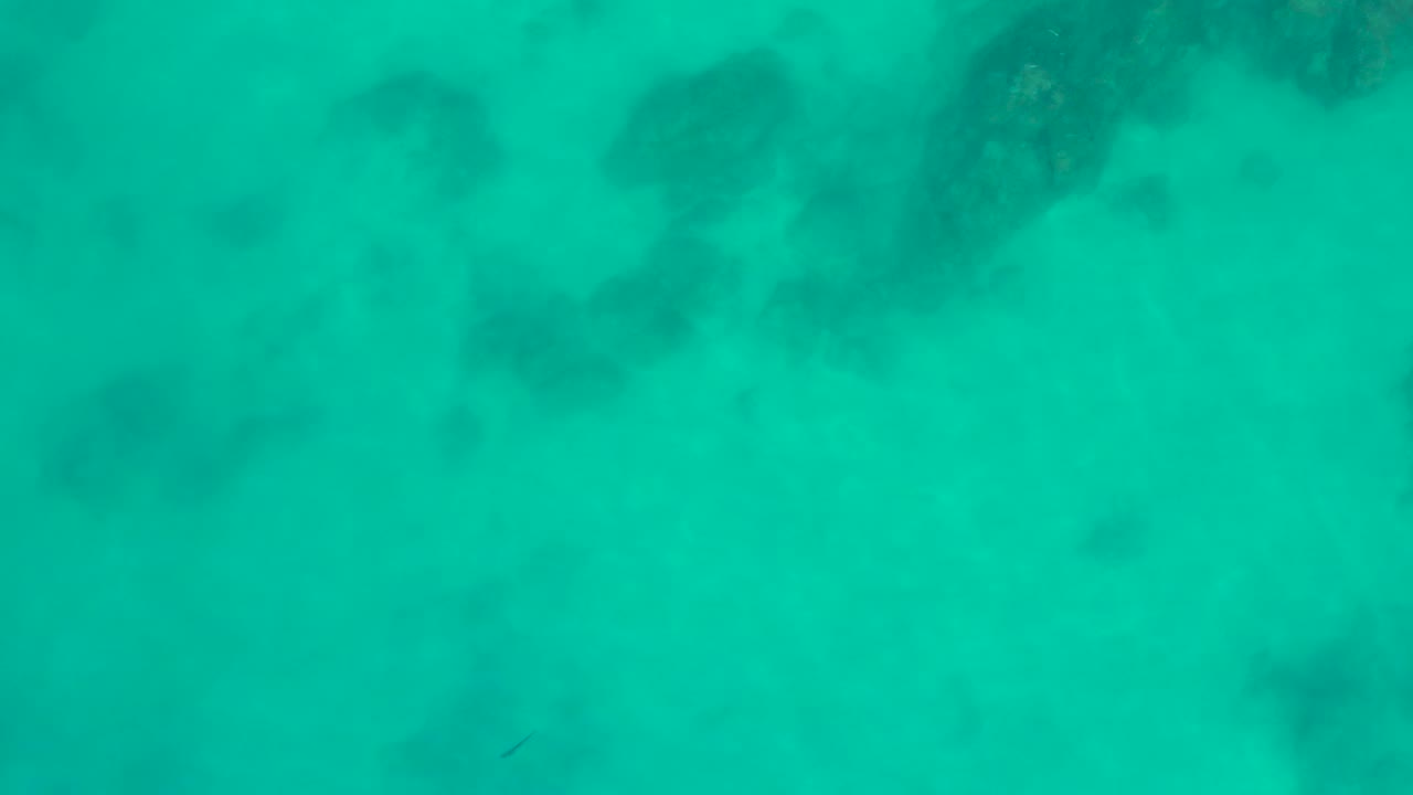 A high overhead frame shows pale seabed under turquoise water with darker coral or rock patches at Shipwrecks Los Cabos, Mexico, and no visible people.