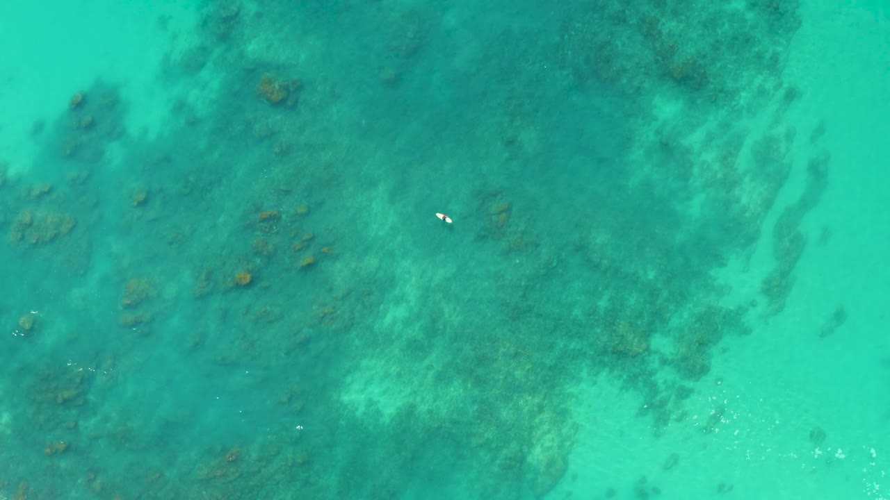 Top-down Shipwrecks Los Cabos frame with mottled reef shapes under turquoise water and the swimmer appearing as a small white mark in the upper area.