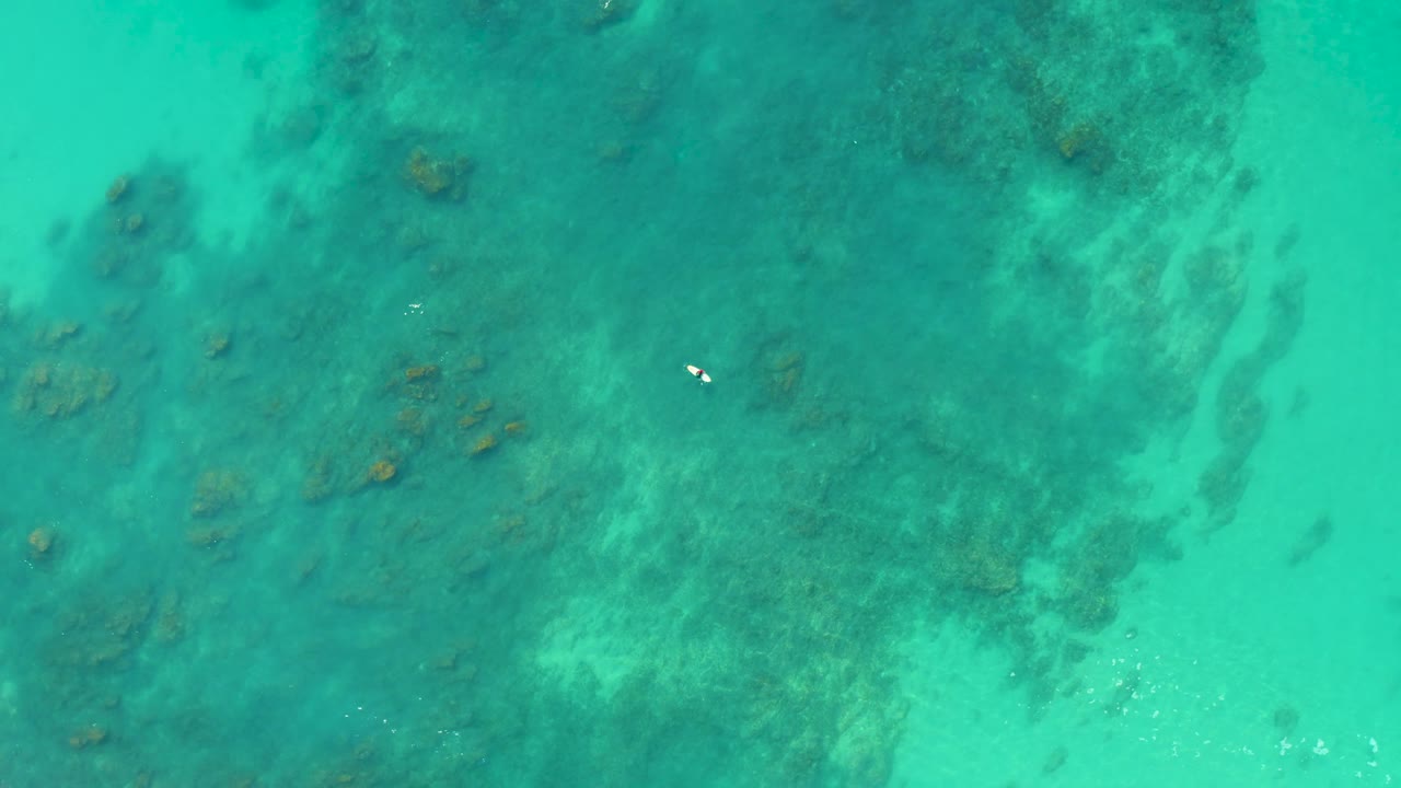 Aerial frame over Shipwrecks Los Cabos shallow reef water, with pale sand channels cutting through darker coral patches and the swimmer still distant.