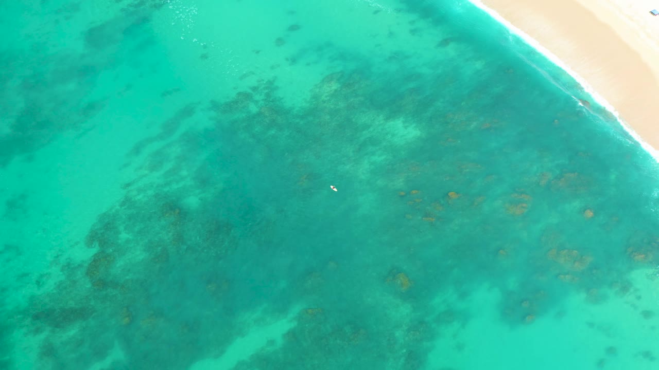 Overhead view at Shipwrecks Los Cabos showing turquoise shallow water, dark underwater patches, a sandy beach edge at upper right, and a tiny white object in the water.