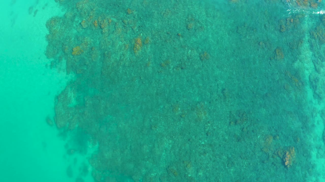 Clear turquoise water covers a dark reef field at Shipwrecks Los Cabos, with small tan formations visible through the surface and no visible people.