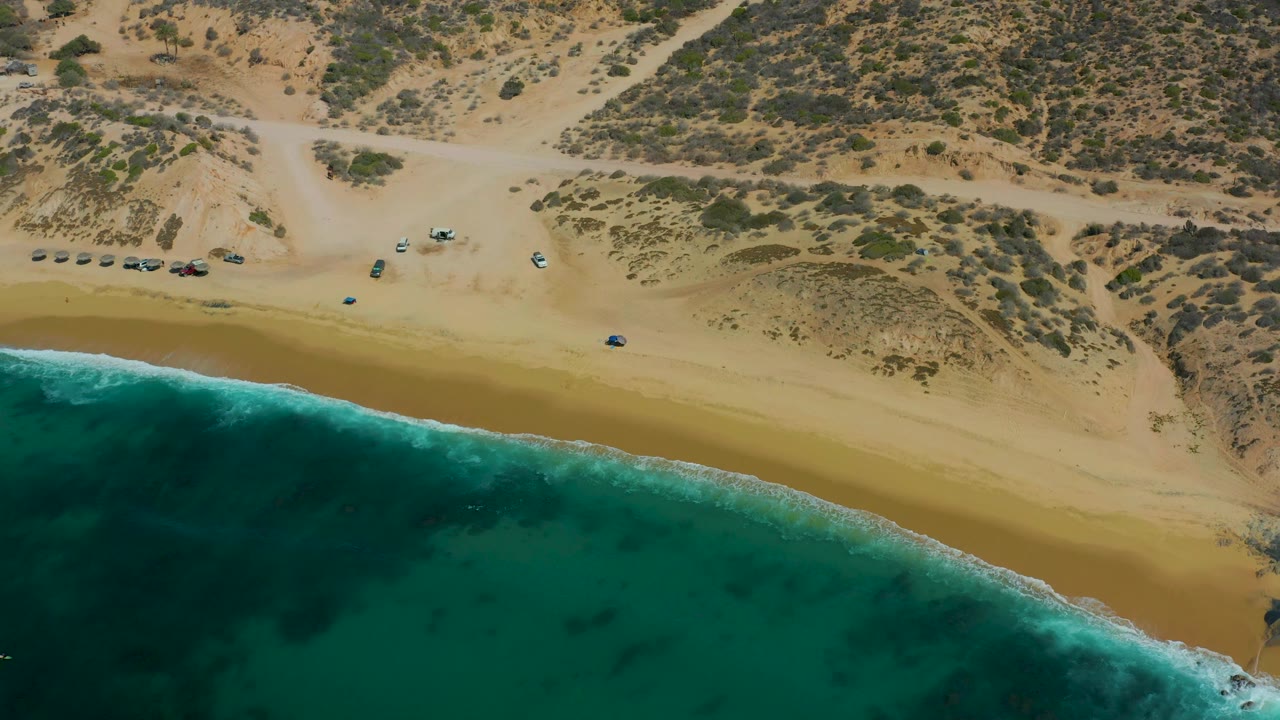 The second frame keeps the Shipwrecks Los Cabos shoreline centered, with vehicles spaced along the upper sand and a darker turquoise patch of water below, with no visible people.