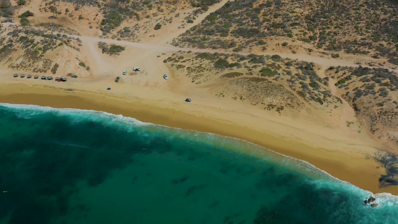 The 0:03 frame shows turquoise water across the lower left, a smooth tan beach band, multiple vehicles near sandy tracks, and scrub-covered dunes at Shipwrecks Los Cabos, with no visible people.