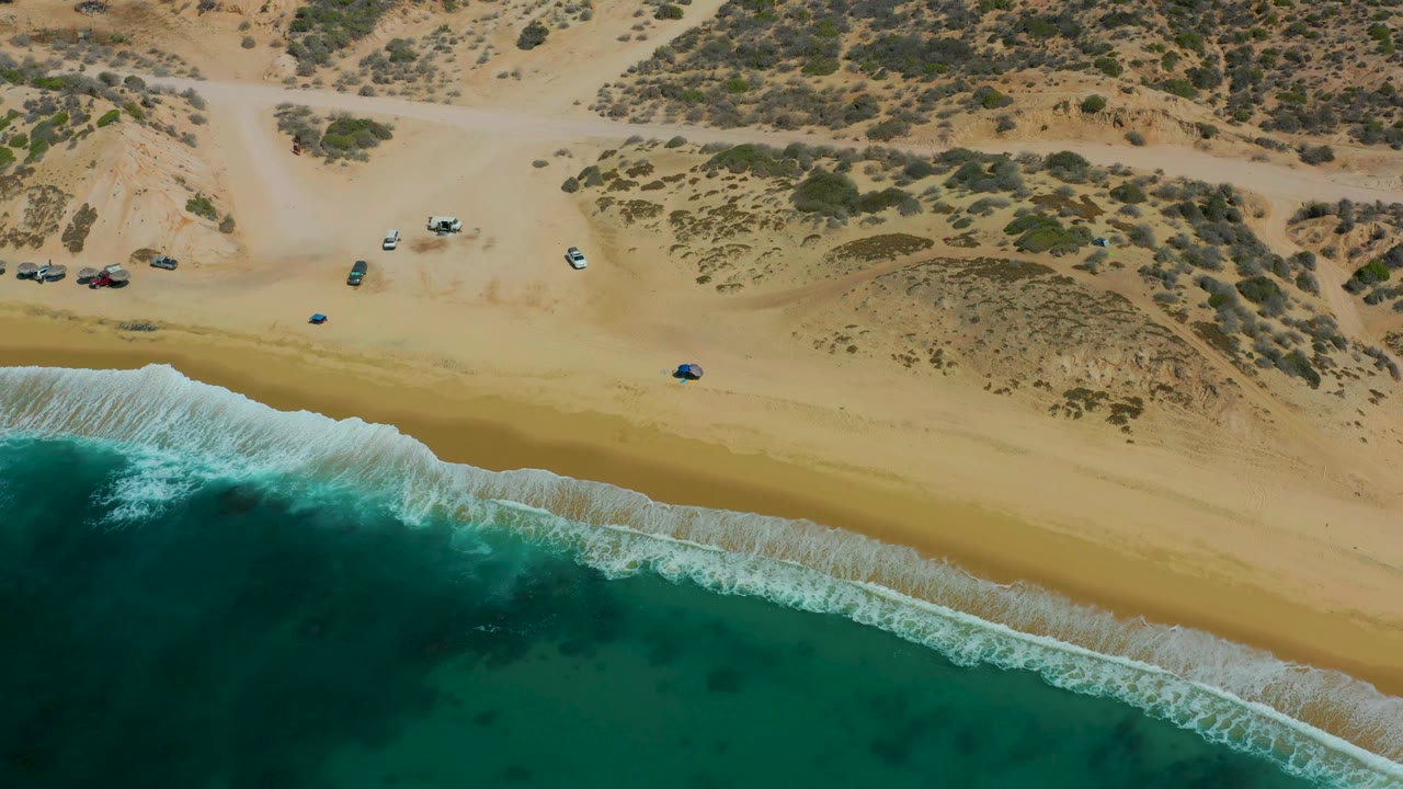 Aerial frame at Shipwrecks Los Cabos with a broad strip of sand, breaking white waves, a parallel dirt road, dune scrub, parked vehicles, and no visible people.