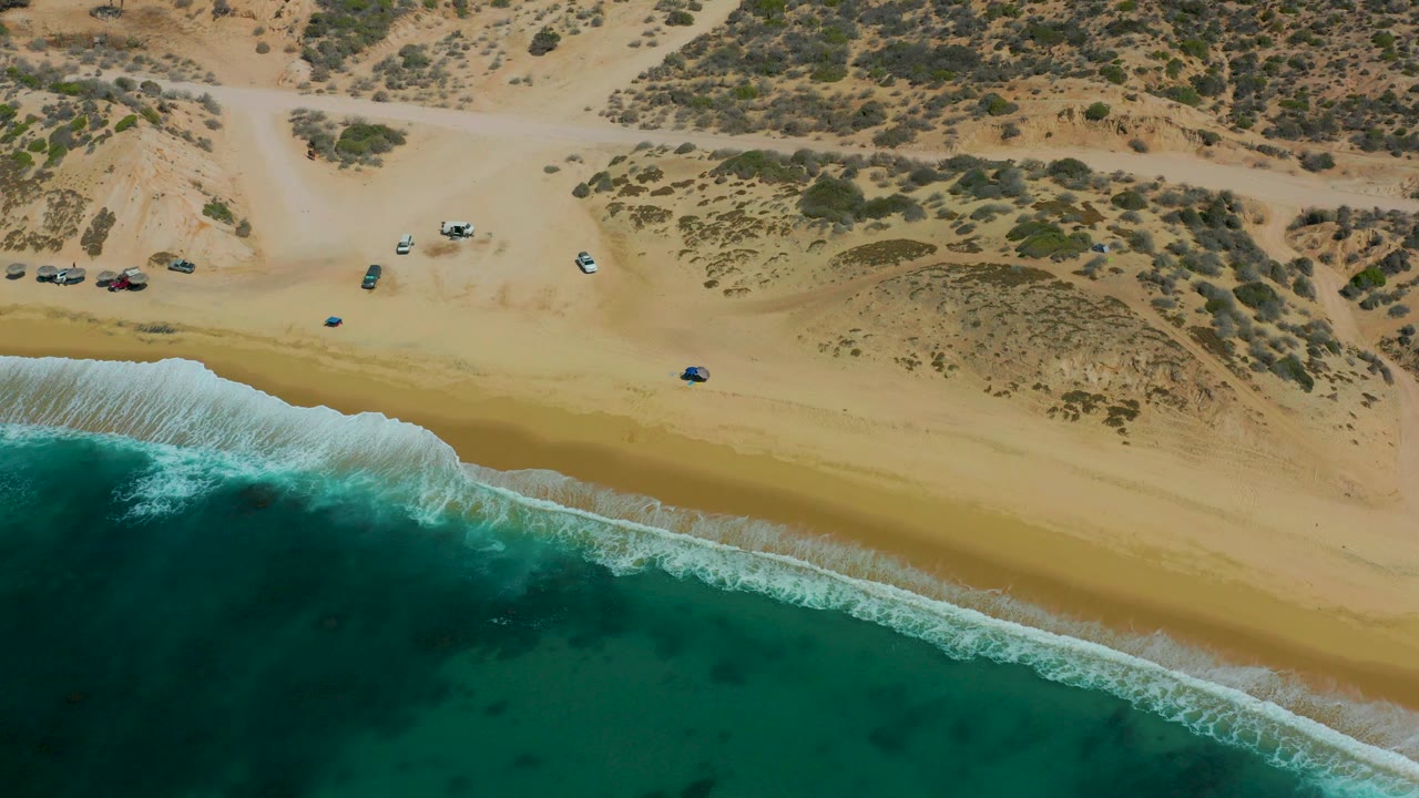 Drone view over Shipwrecks Los Cabos with turquoise ocean in the lower frame, a clean sand band, dune shrubs and vehicle tracks above, parked vehicles, and no visible people.