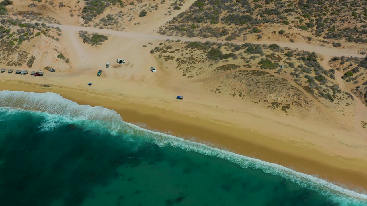 Wide overhead beach scene with surf, dunes, road, and small parked cars.
