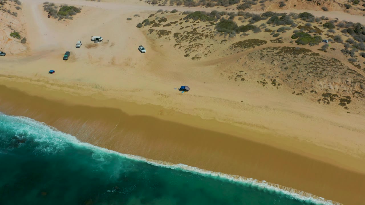 Aerial frame at Shipwrecks Los Cabos with the surf line running diagonally across the lower beach, several vehicles spread across the sand, scrub-covered dunes above, and no visible people.