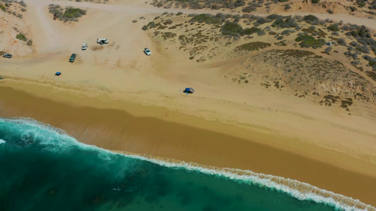 High drone view over Shipwrecks Los Cabos with white surf entering from the lower left, a wide empty strip of sand, vehicles near the upper beach, dune vegetation, and no visible people.