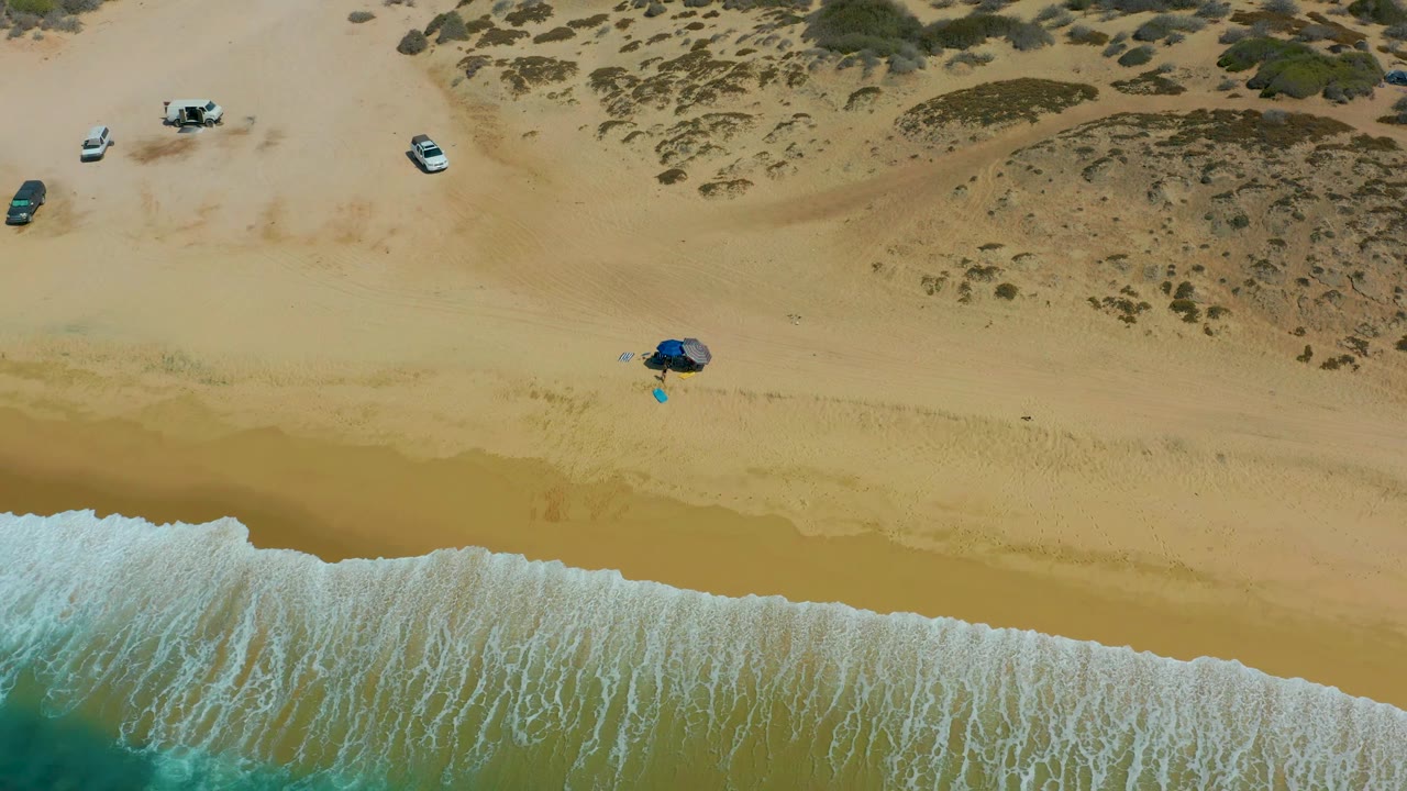 At 0:01, the Shipwrecks Los Cabos shoreline shows a wider band of white foam over wet sand, with the blue umbrella near center beach, vehicles higher on the sand, and no visible people.