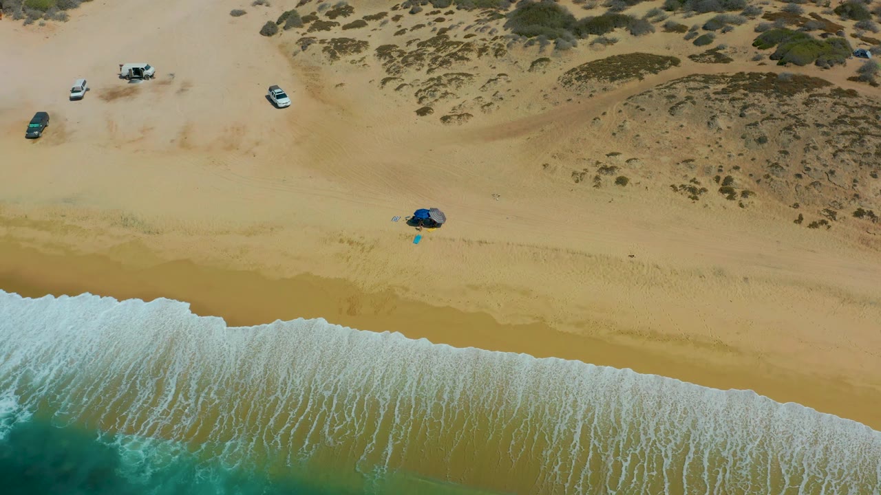 At 0:02, white surf forms a continuous bright edge across the lower Shipwrecks Los Cabos beach, with tan sand, scattered parked vehicles, dune scrub, and no visible people.