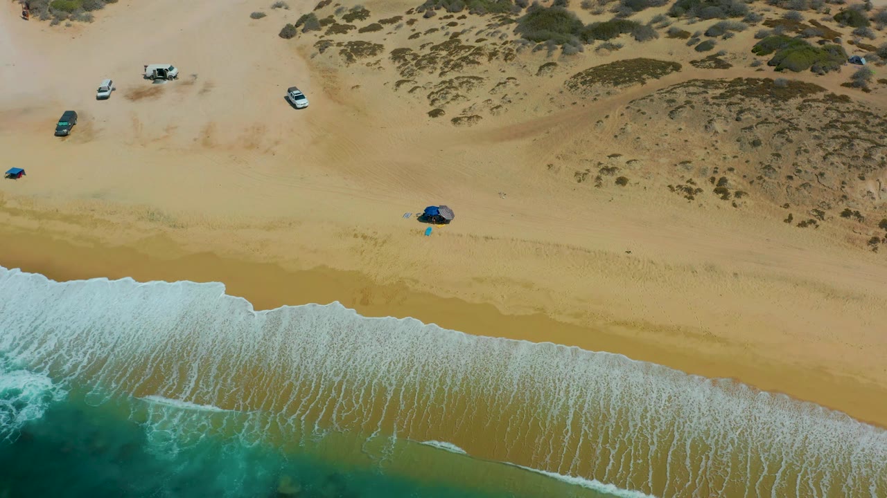 At 0:03, the aerial view over Shipwrecks Los Cabos places foamy water at the lower left, open sand through the center, parked cars near the upper sand, shrubs on dunes, and no visible people.