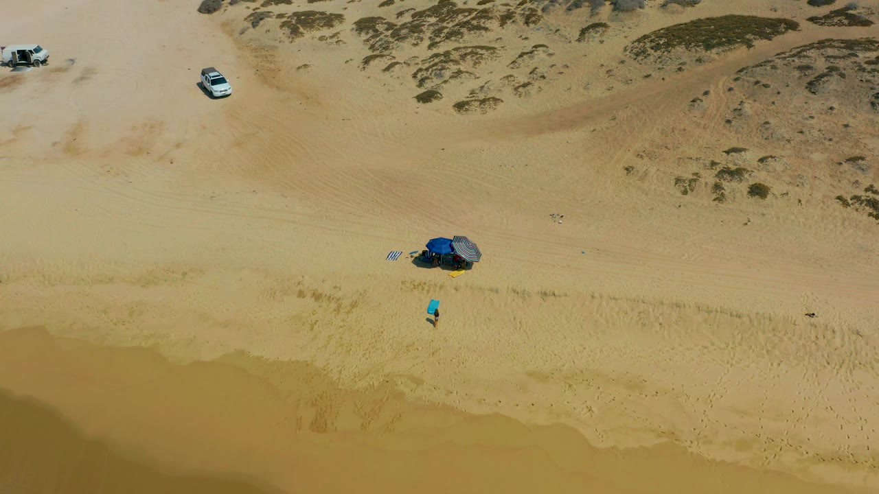 Overhead view of Shipwrecks Los Cabos beach with tan sand, scrub-covered dunes, two visible vehicles, a blue tent setup near the waterline, and a small person-sized figure nearby.