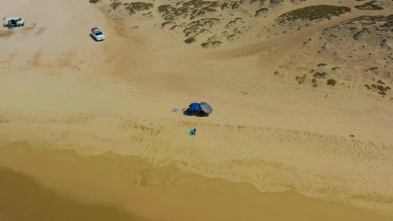 High downward view of Shipwrecks Los Cabos beach with the blue tent setup slightly right of center, a small person-sized figure below it, vehicles near the dunes, and wet sand along the lower edge.