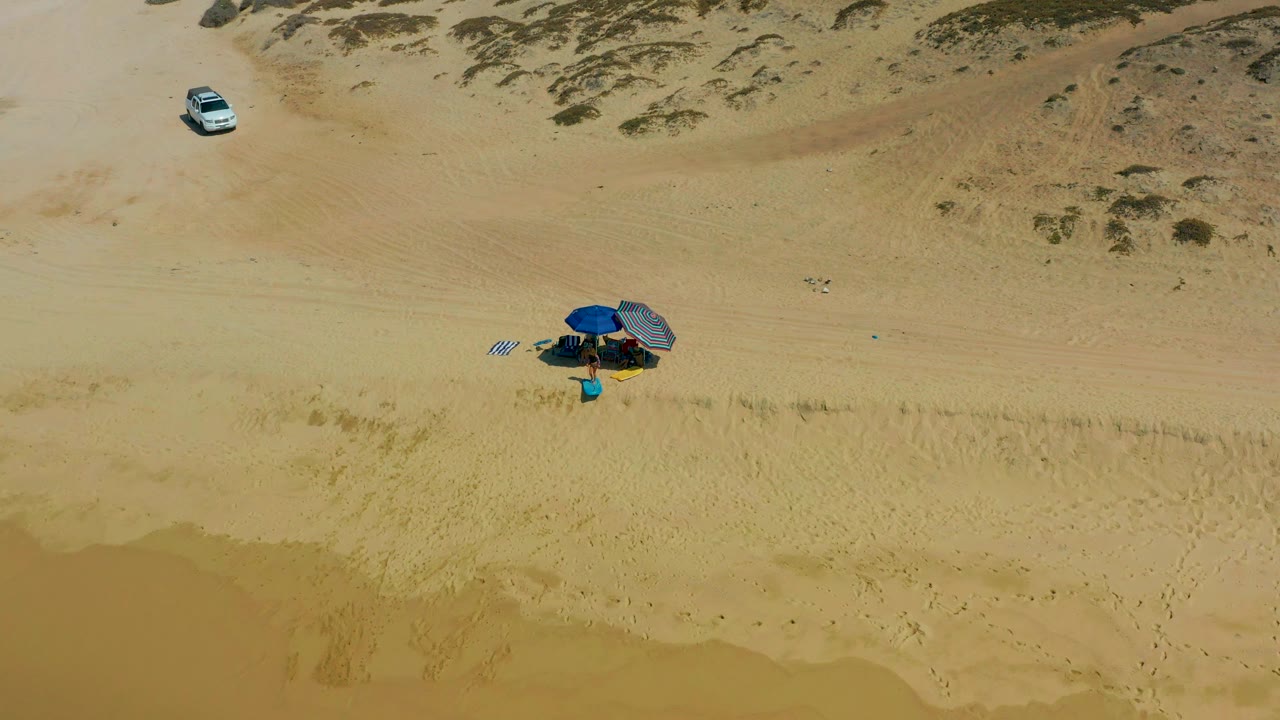 Aerial frame from Shipwrecks Los Cabos showing a blue umbrella and beach gear on open sand, a white vehicle near the upper dunes, and shallow water along the lower edge.