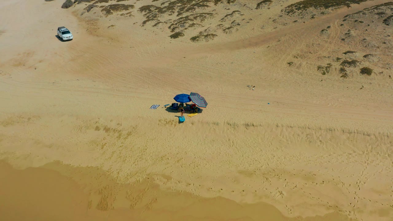 Blue umbrella and beach gear on sand with a vehicle near dunes.