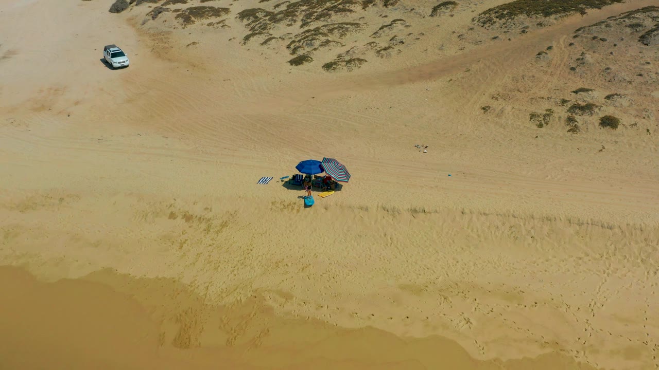 High drone frame at Shipwrecks Los Cabos showing the umbrella area on empty sand, a white vehicle above it, dune scrub in the background, and the wet shoreline low in the frame.