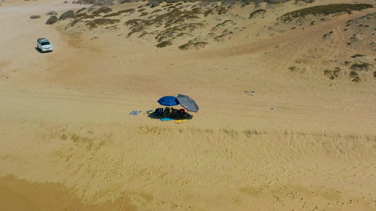 The white SUV has moved farther across the open sand above the blue umbrella and striped shade at the Shipwrecks Los Cabos campsite, with no visible people.