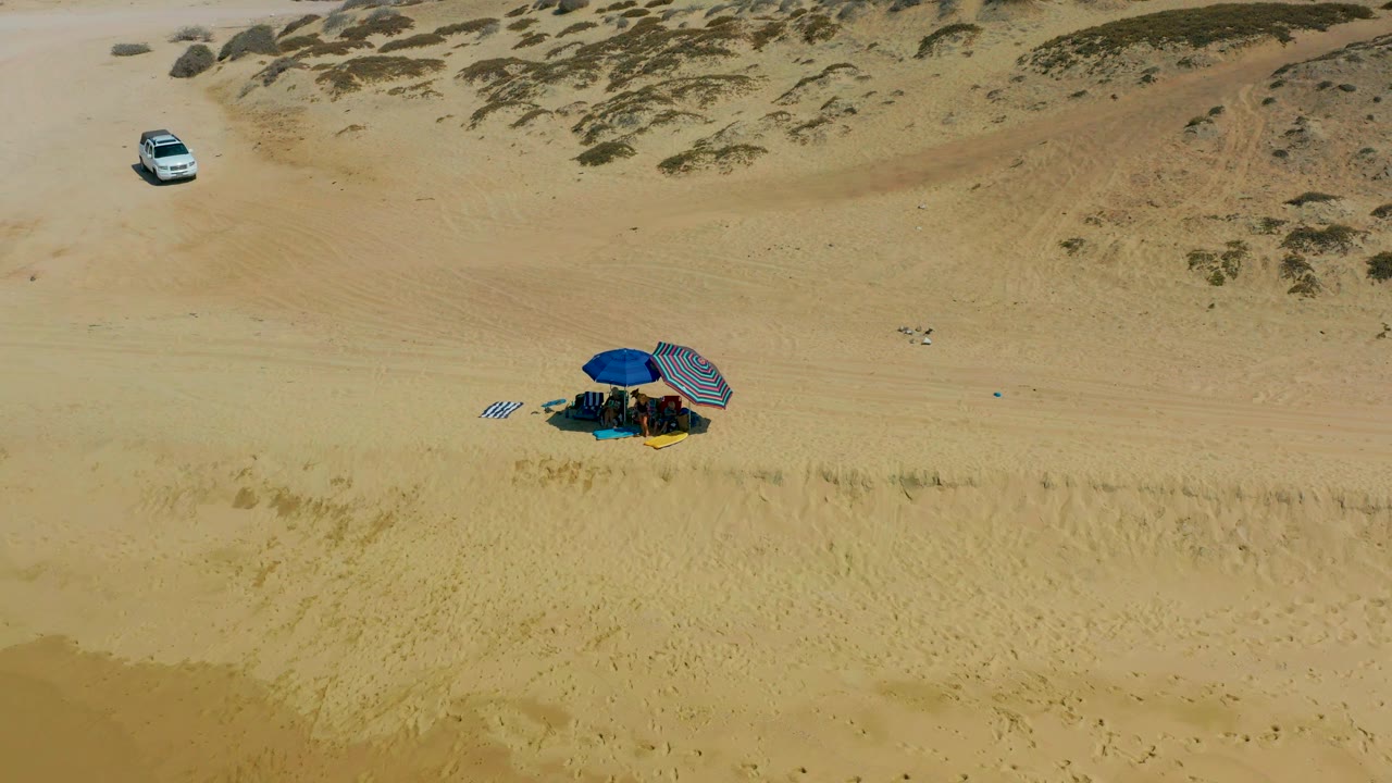 The SUV sits higher in the frame near scrub-covered dunes while the blue umbrella campsite remains below on pale sand at Shipwrecks Los Cabos, with no visible people.