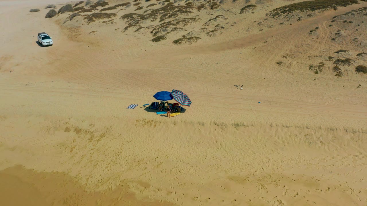 Midway through the clip, the aerial view holds the white SUV in the upper left area and the blue umbrella camp lower in frame on sandy Shipwrecks Los Cabos terrain, with no visible people.