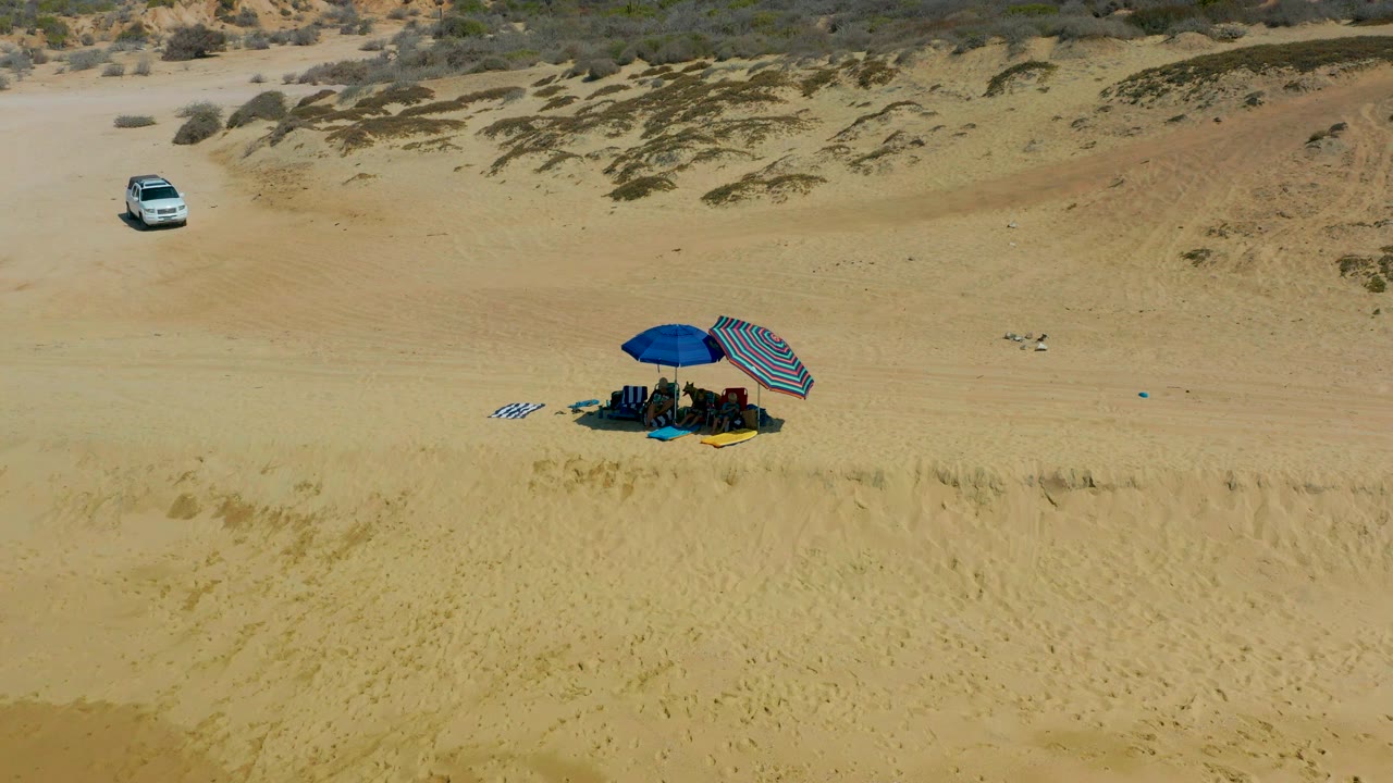Aerial view at Shipwrecks Los Cabos showing a white SUV on upper sand and a small beach camp with blue and striped umbrellas below.