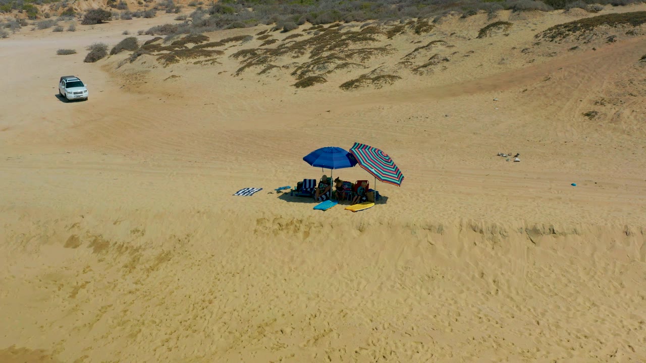 At 0:02, the aerial view holds on the umbrella group at Shipwrecks Los Cabos, with bright sand around them and a white car set back near the scrub-covered dunes.
