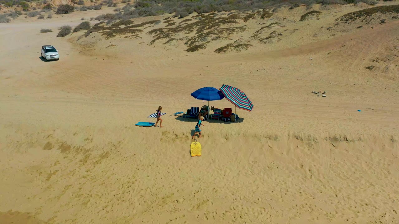 At Shipwrecks Los Cabos, an aerial frame shows a wide sandy beach, a small umbrella setup, blue and yellow surfboards, visible people, and a white car parked near the dunes.