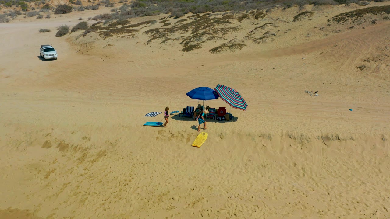 At Shipwrecks Los Cabos, visible people remain beside the umbrellas while the yellow surfboard sits in the lower midground and the white car stays parked on the sandy track behind them.