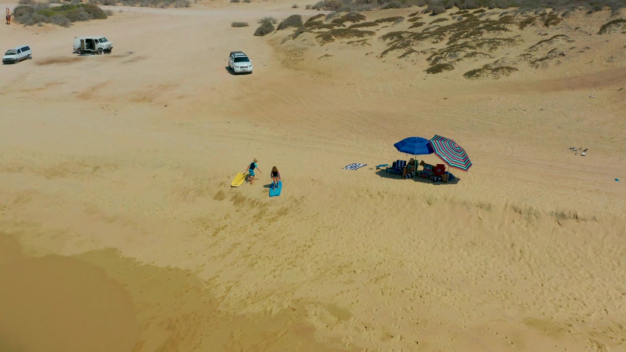 Two people with a yellow surfboard walk across sand toward umbrellas in a wide aerial beach view.