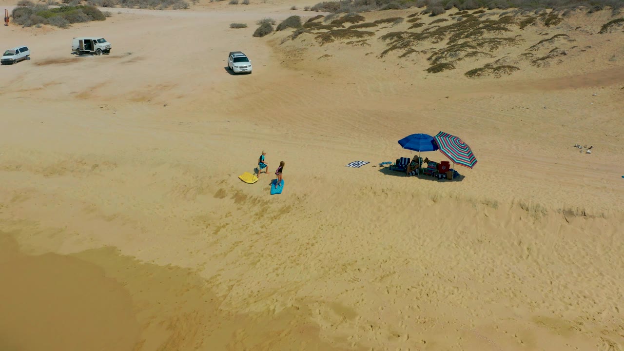 At Shipwrecks Los Cabos, the two people continue across the dune slope with the yellow surfboard, while the blue and striped umbrellas sit to the right and white vehicles remain in the distance.