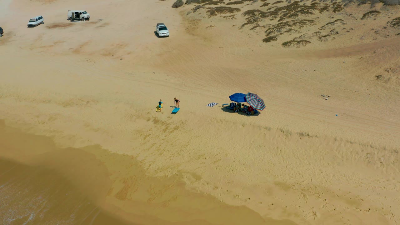 At Shipwrecks Los Cabos, two small beachgoers stand near towels and a blue umbrella on open sand, with several vehicles parked near low dunes in the background.