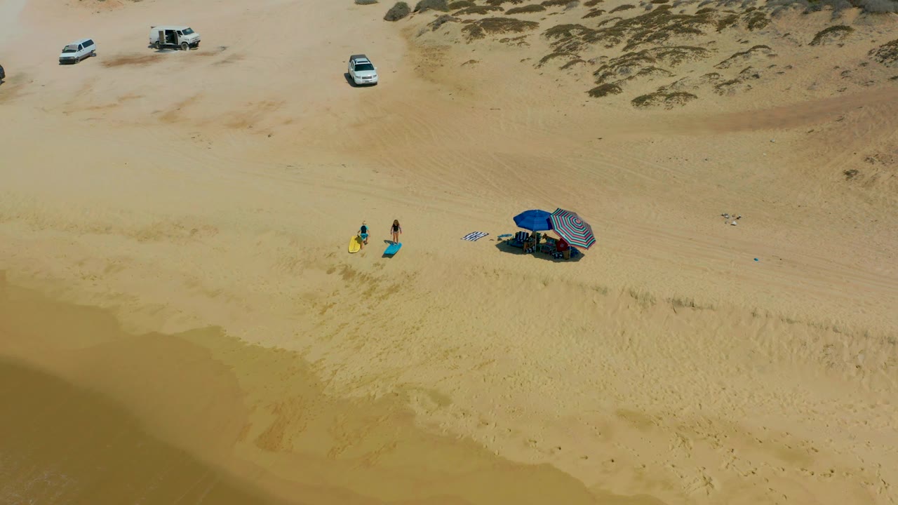 At Shipwrecks Los Cabos, two beachgoers are separated slightly on the sand near the umbrella setup, with cars above them and shallow water along the lower edge.