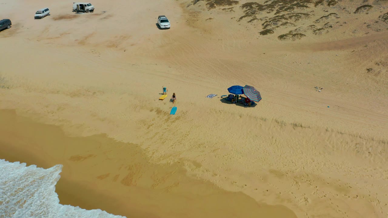 Aerial frame at Shipwrecks Los Cabos where a larger wedge of white surf reaches in from the lower left, with two beachgoers, colored boards, umbrella shade, and parked cars remaining spread across the sand.