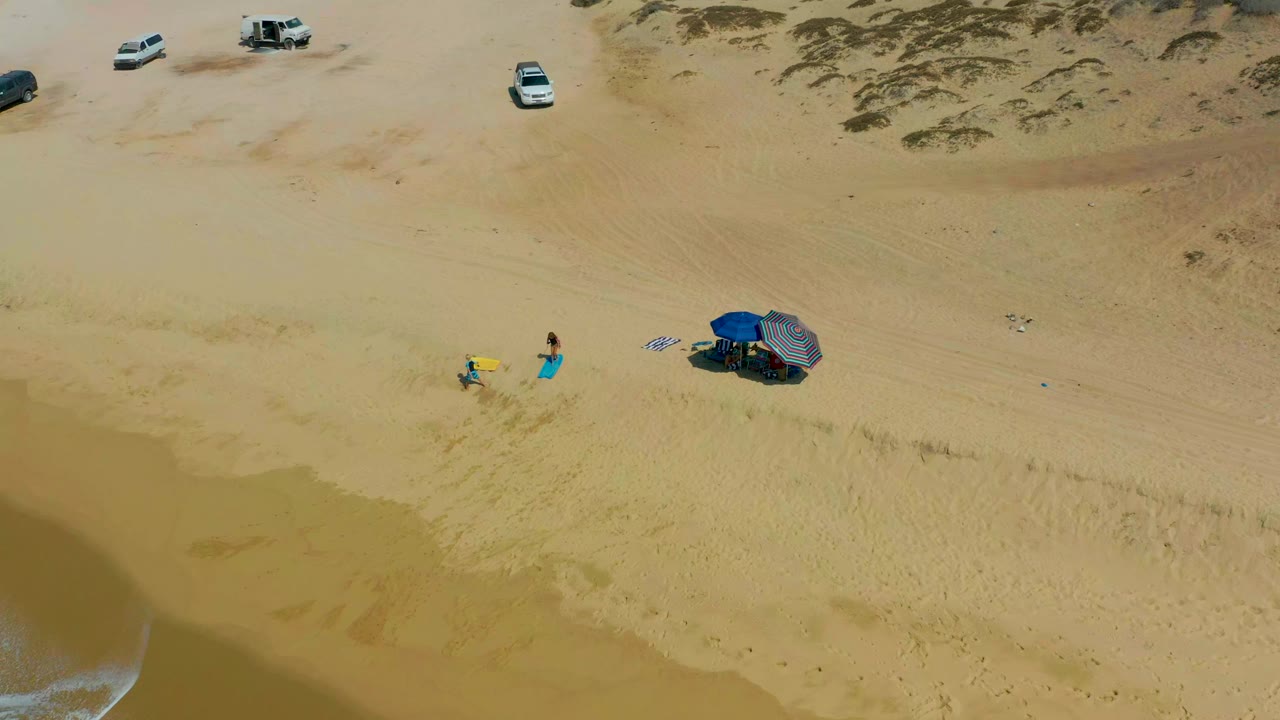 Overhead frame from Shipwrecks Los Cabos showing two people near beach gear on tan sand, umbrellas to the right, parked vehicles near the dunes, and surf entering at the lower left.