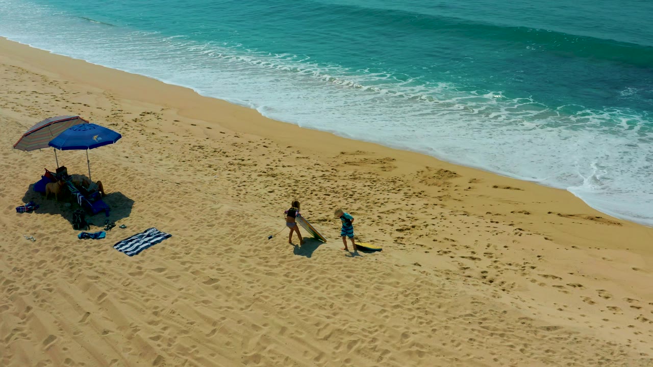Aerial view at Shipwrecks Los Cabos showing two people near a blue umbrella, towels, beach gear, a yellow surfboard, sandy footprints, and turquoise surf.