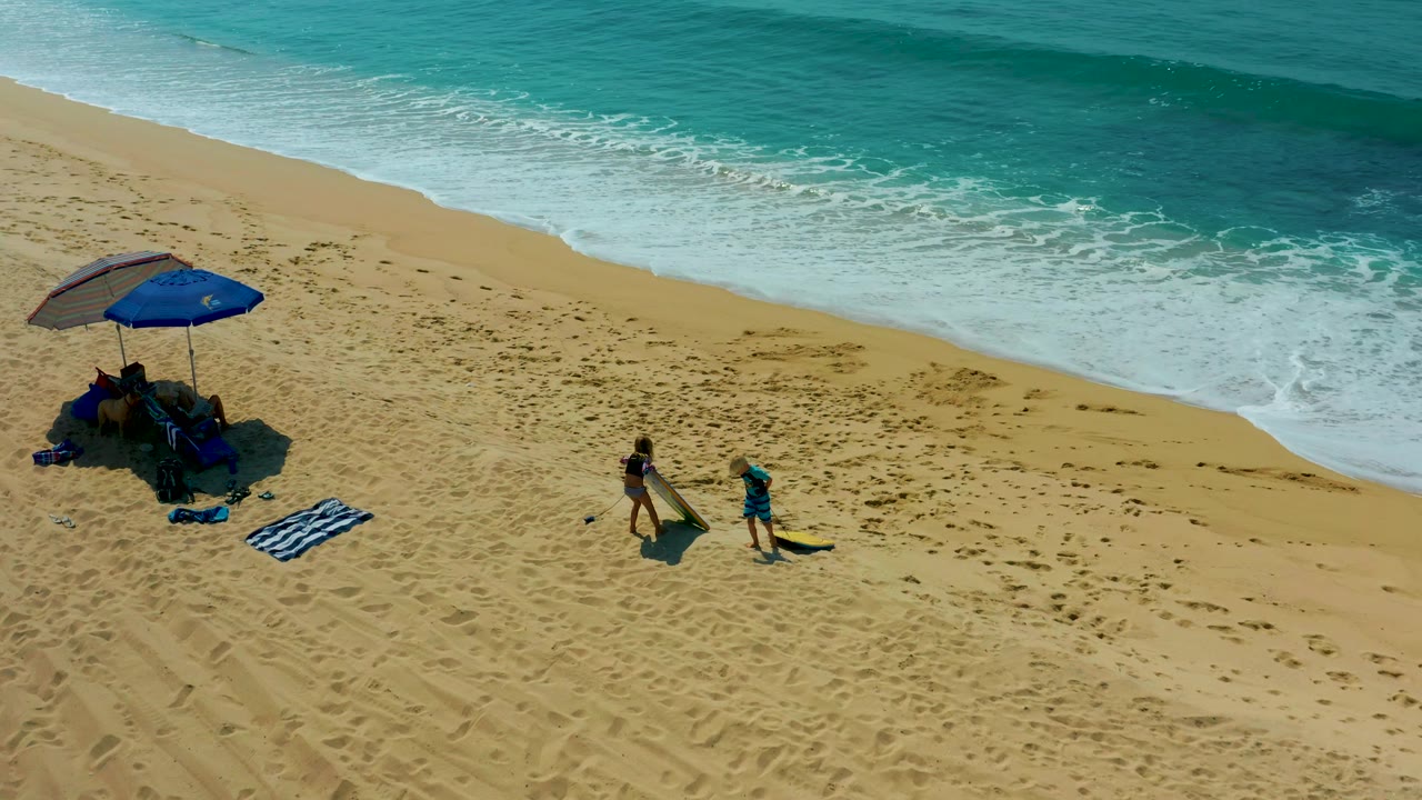 At Shipwrecks Los Cabos, two people remain visible on the sandy shore near a blue umbrella while white foam advances along the upper edge of the frame.