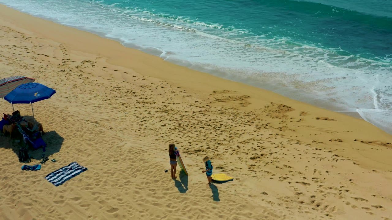 A high aerial frame at Shipwrecks Los Cabos shows the blue umbrella close to the left edge, two people farther right, a yellow surfboard, footprints, and breaking waves above them.