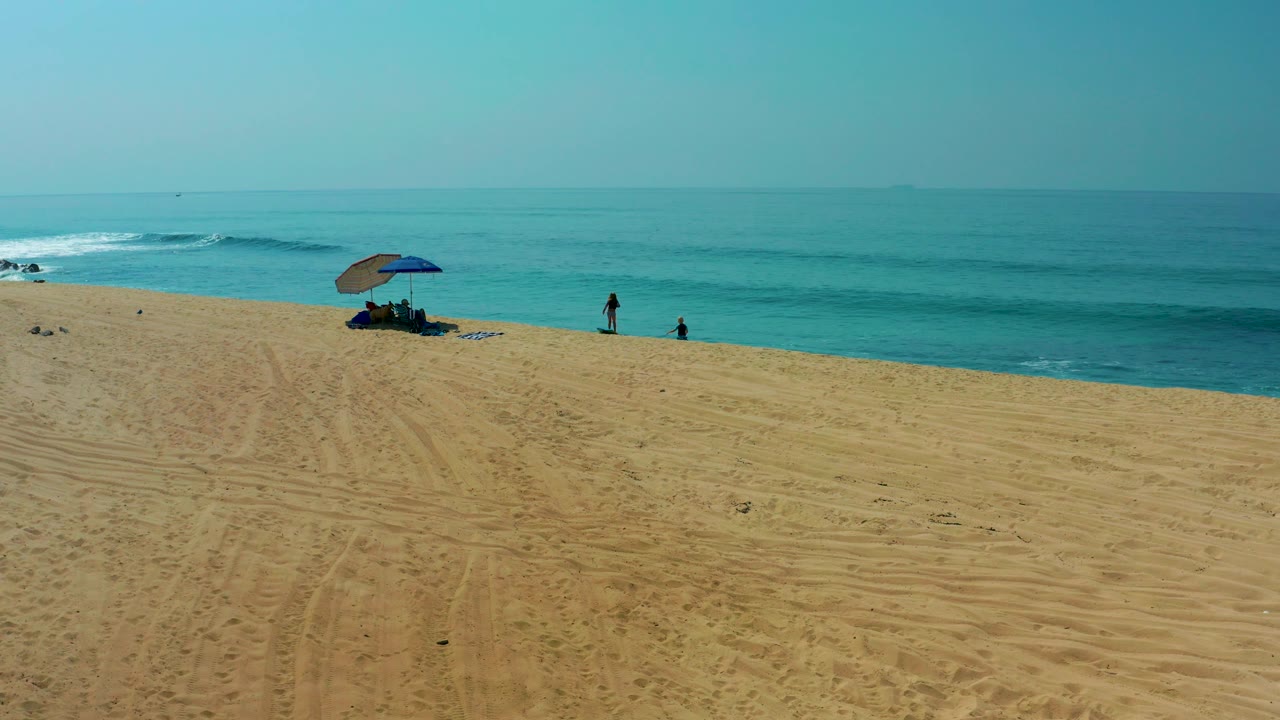 Shipwrecks Los Cabos beach view with tire-marked sand in front, a shaded umbrella near the waterline, small beachgoers, and low turquoise surf offshore.